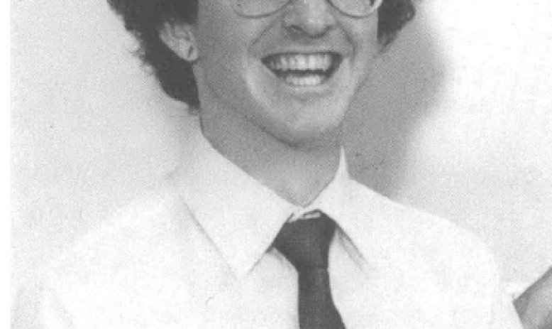 Photo of white man with glasses in collared shirt and tie grinning at the camera 