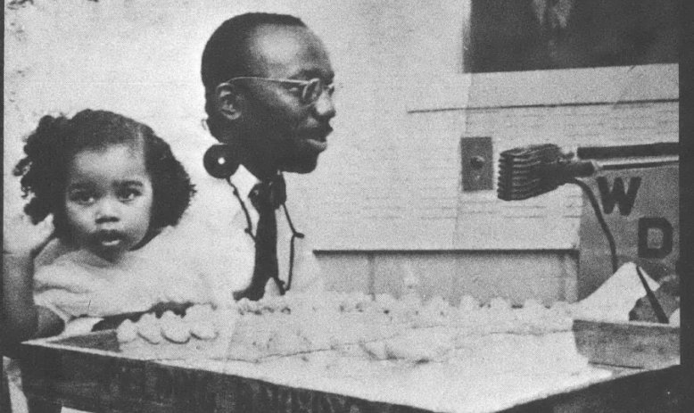 Black and white photo of Black man with glasses seated at table speaking into microphone, with toddler-age child seated next to him looking away from the camera