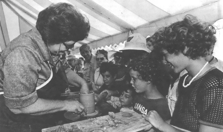 Black and white photo of older white woman showing a piece of pottery to children at a fair booth