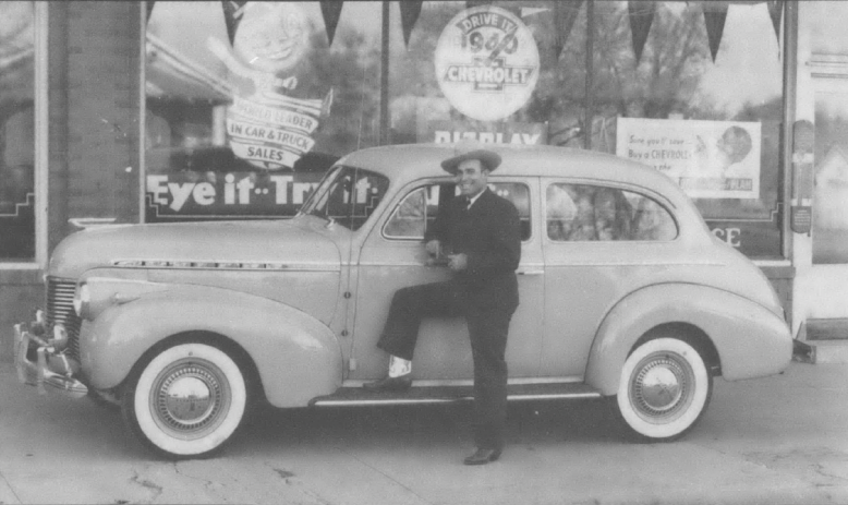 Black and white photo of man standing next to nice car in front of shop window