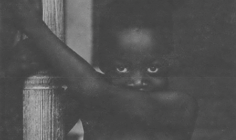 Shirtless Black child holding on to a wooden post and peering over their arm