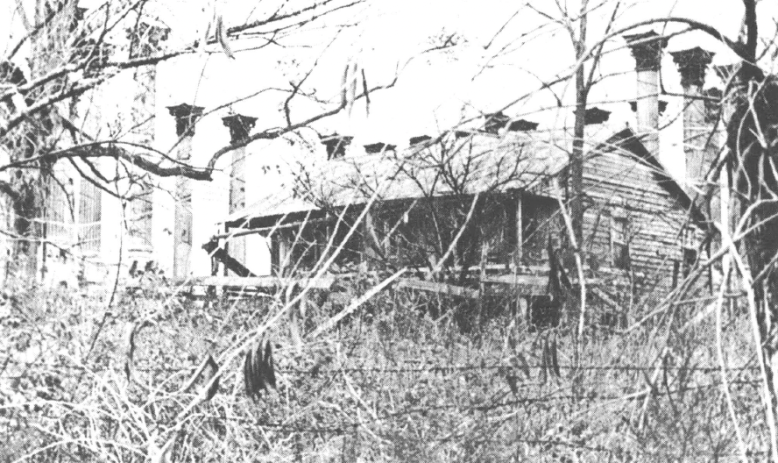 Black and white photo of building behind a barbed wire fence and tall grass