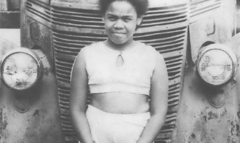 Black and white photo of Black child standing in front of old car