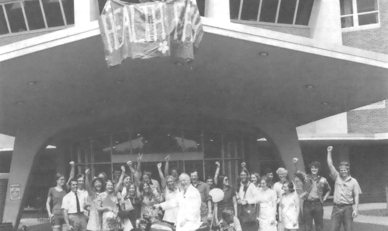 Black and white photo of group of people with fists raised standing in front of a building. A person standing on a balcony holds a banner