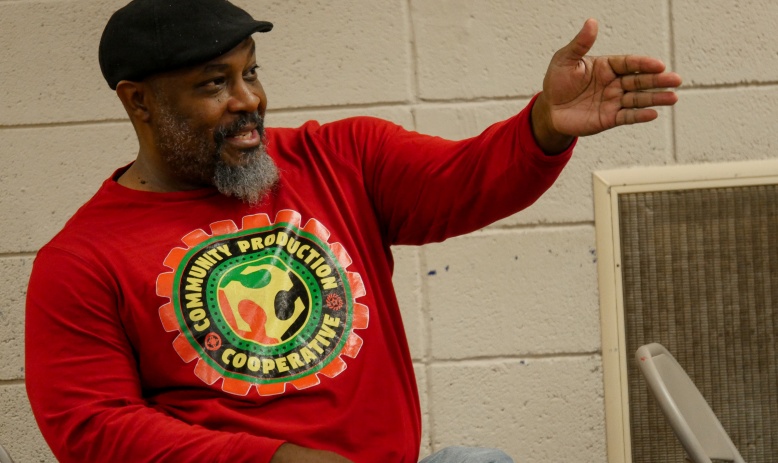 A man seated in front of a cinderblock wall wearing a black cap, red long-sleeve t-shirt, and jeans, gestures while talking