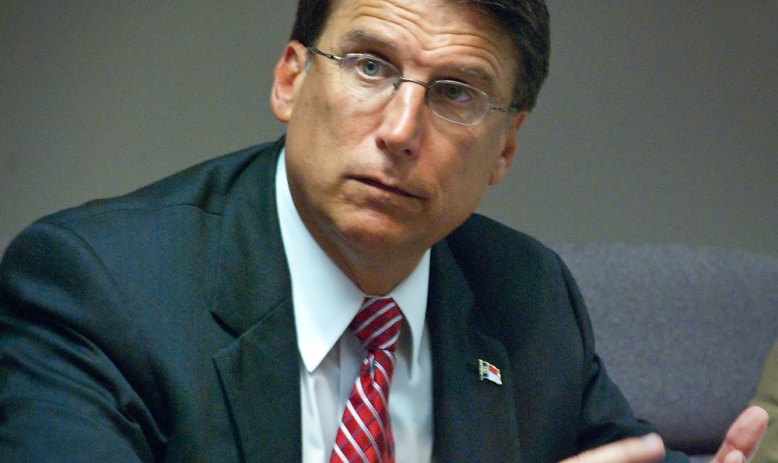 The photo depicts a white man with glasses and brown hair counting on his fingers while looking outside of the camera's view to the upper lefthand corner of the image. He is wearing a dark-colored suit, blue dress shirt, and red tie.