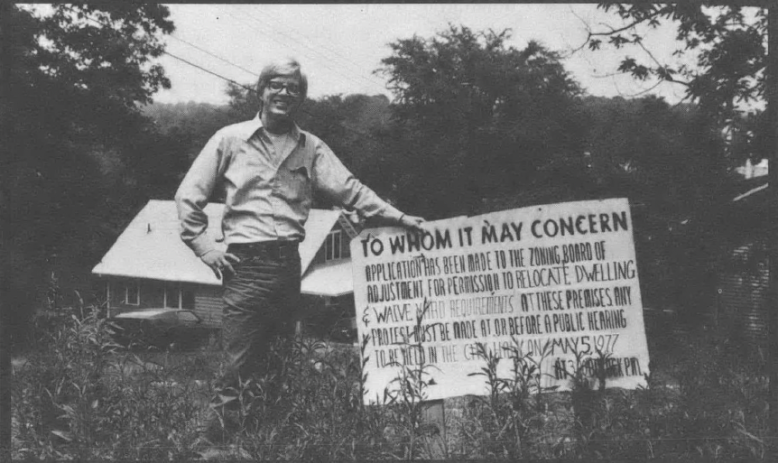 Black and white photo of white man standing in front of sign in yard, in front of house