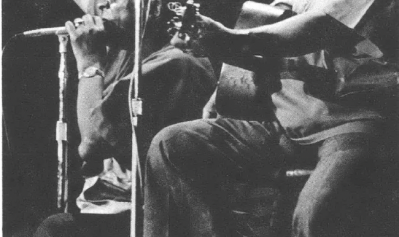 Black and white photo of two Black men, one sitting in front of a microphone playing the harmonica and the second sitting in front of a microphone smiling and playing guitar 