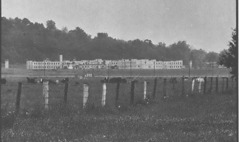 Black and white photo of building far from camera, with a large field in front and a fence