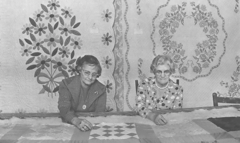 Two old white women sitting at a table with quilts in front of two quilts hanging on the wall