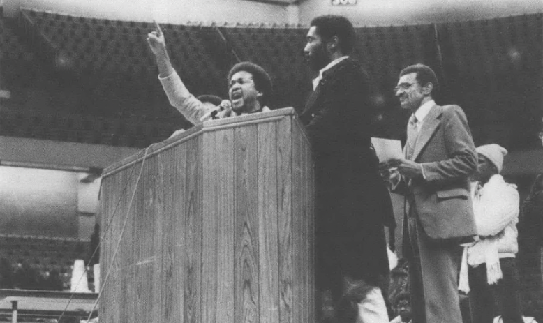 Black and white photo of three men speaking behind podium, one with arm raised and finger pointed up