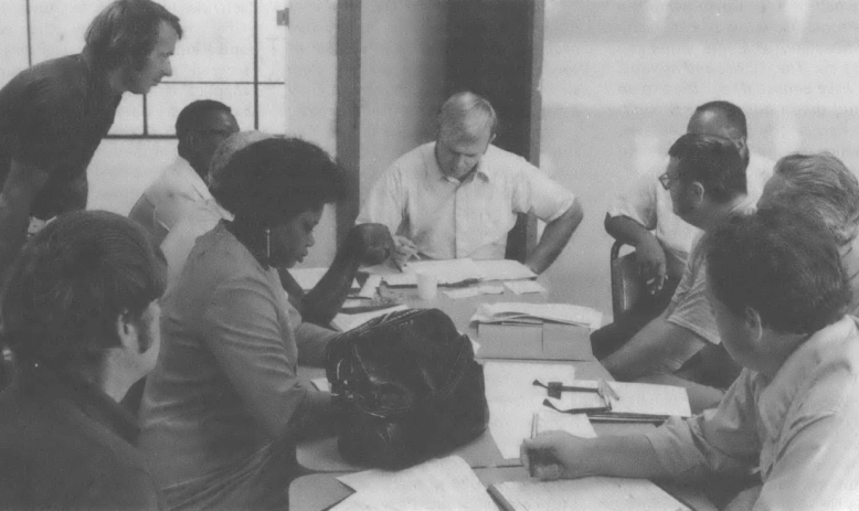 Black and white photo of multiracial, multi-gendered group of people sitting around a table having a meeting
