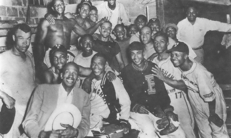 Black and white photo of roughly twenty Black men in dugout or locker room smiling at camera