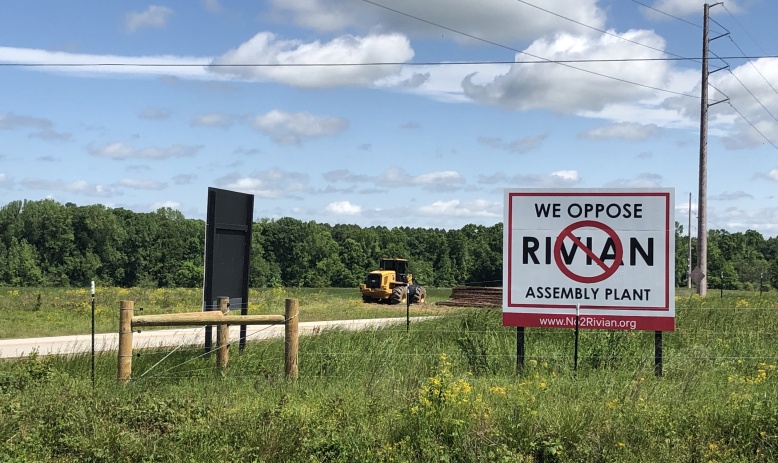 The image shows a field with a yellow bulldozer in the background and a white and red sign in the foreground that reads, "We Oppose Rivian Assembly Plant." The field is also offset by a blue sky with white clouds.