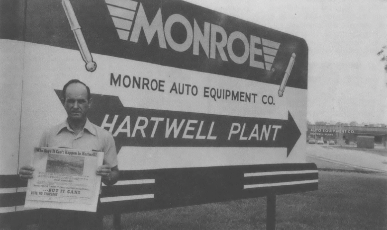 Black and white photo of middle aged white man standing in front of billboard advertising auto manufacturing plant