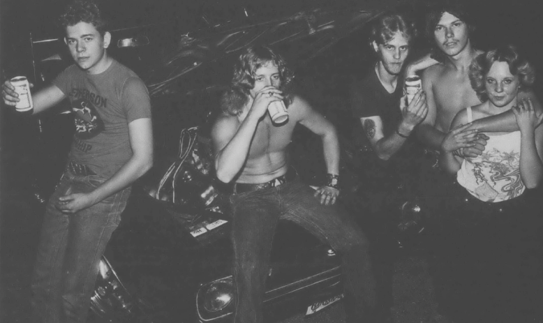 Black and white photo of white teenagers sitting on car hoods and drinking something out of cans