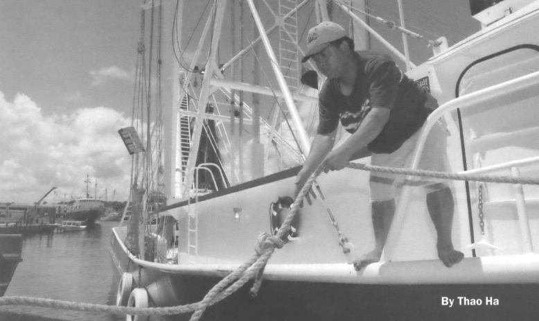 Black and white photo of a woman standing on the side of a boat pulling a rope out of the water. Text in the bottom right corner reads "Troubled Water by Thao Ha"