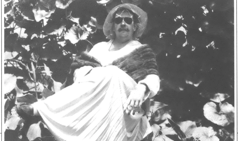 Black and white photo of man wearing sunglasses and floppy hat and dress sitting under shade tree