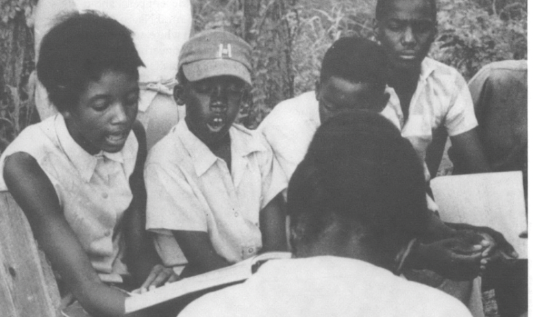 A group of Black students seated outside reading a book. One appears to be reading out loud.