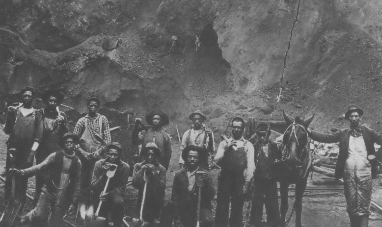 Black and white photo of about a dozen Black men in work clothes posing with tools