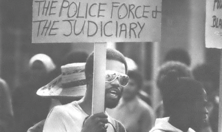 Black and white photo of Black man holding sign that reads "Remove all Klansmen from the police force & the judiciary"