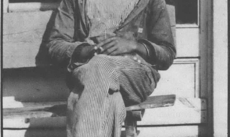 Photo of a mustachioed Black man sitting on a wooden bench, wearing bibbed overalls, a work jacket, and a cap with a brim
