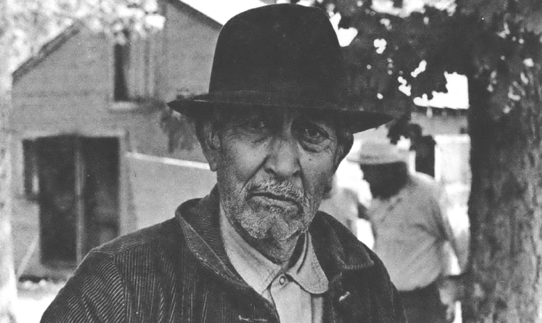 Black and white headshot of older man with bowler hat and wearing a collared shirt and jacket standing in front of a barn