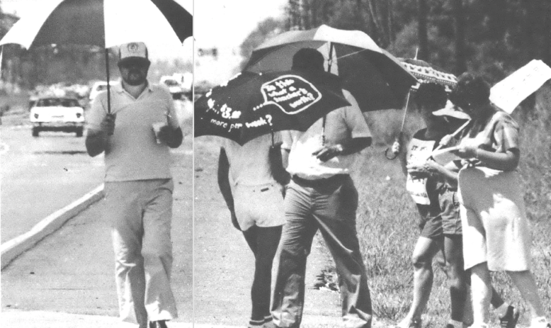 Photo of several people picketing on the side of the road with umbrellas for the sun