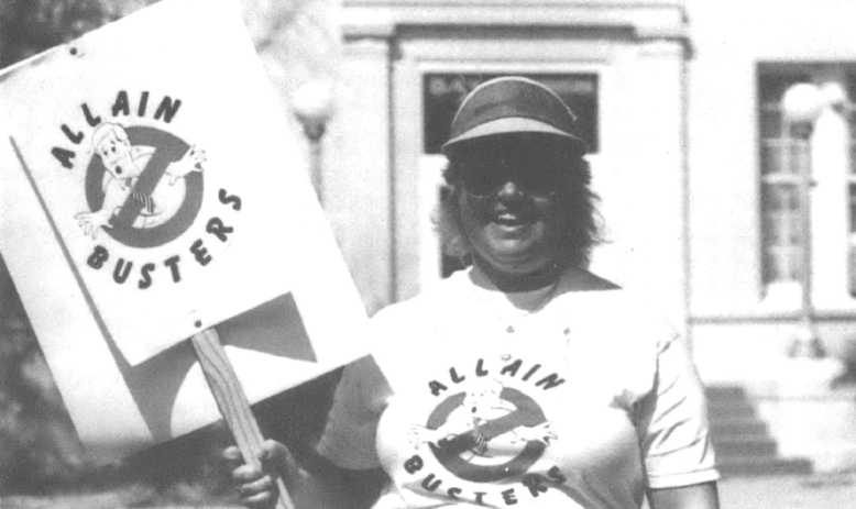 Photo of woman in visor with t-shirt and sign that say "Allain Busters" with a Ghostbusters cartoon. She is smiling at the camera