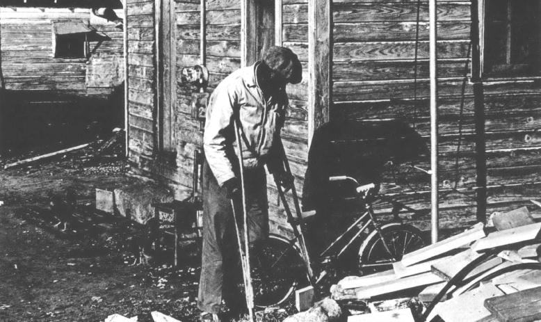 Black and white photo of Black man on crutches in front of a wood home