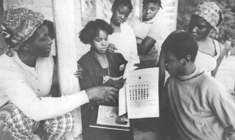 Black and white photo of group of Black children and a Black woman pointing at a piece of paper 