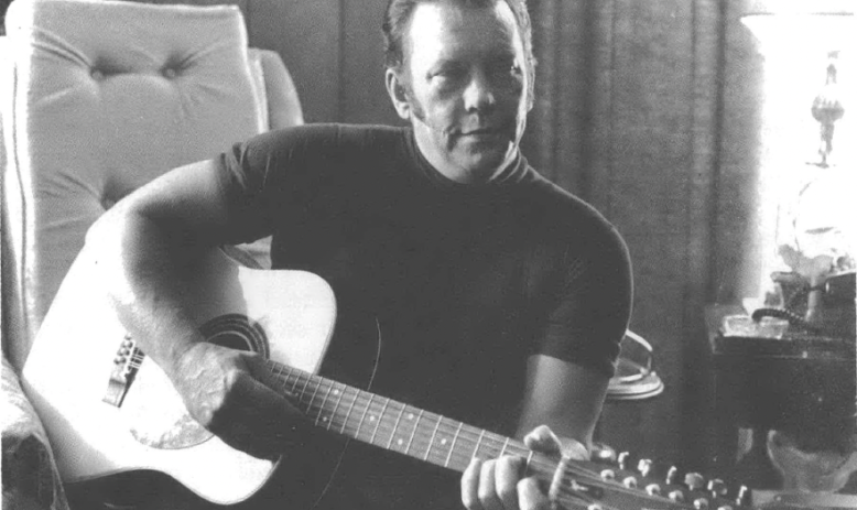 Black and white photo of white man with slicked-back hair, seated and holding a guitar 