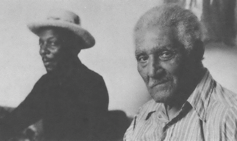 Black and white photo of two Black men, one wearing hat, sitting next to each other looking sideways at the camera