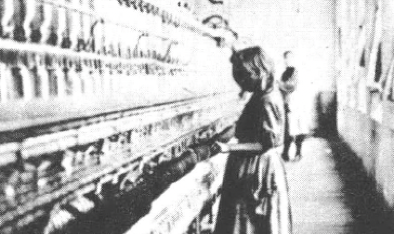 Black and white photo of young girl standing at loom in factory