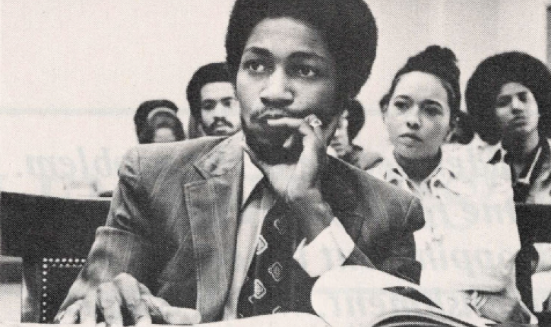 Black and white photo of Black man sitting at desk looking behind camera with papers in front of him