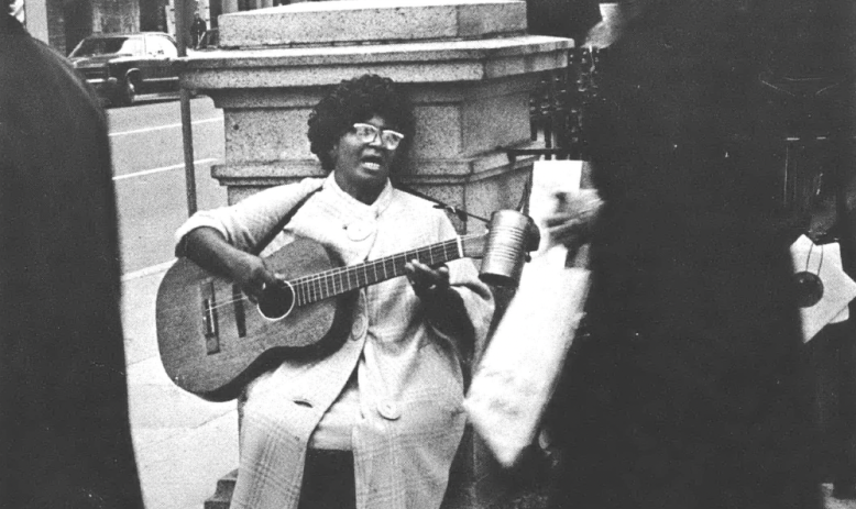 Black and white photo of Black woman in glasses, long coat, singing and playing guitar sitting on a street corner to passersby