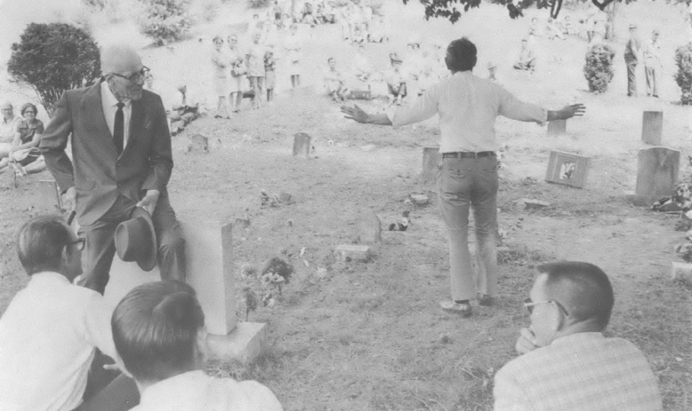 Black and white photo of group of people outside in a graveyard, one man walking away from the camera with his arms out