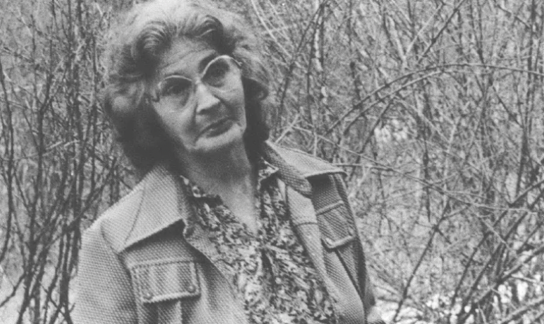 Photo of old woman standing in the woods speaking to someone behind the camera