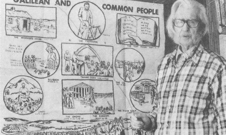 Black and white photo of older white man in glasses and plaid shirt standing in front of a large poster