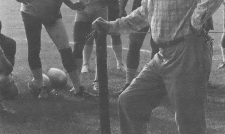 Black and white photo of old man wearing hat standing in front of other men wearing football uniforms