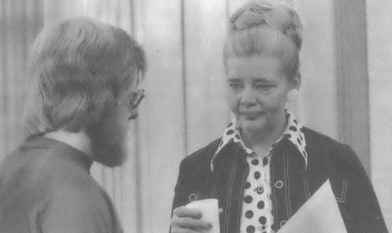 Black and white photo of a younger white man in glasses and beard talking with an older, well-dressed white woman