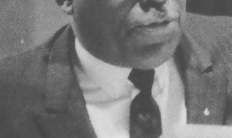 Black and white photo of Black man in suit, wearing glasses, with short white hair giving a speech