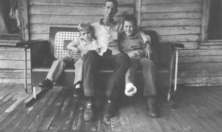 Black and white photo of man holding two children sitting on a bench on the porch of a house