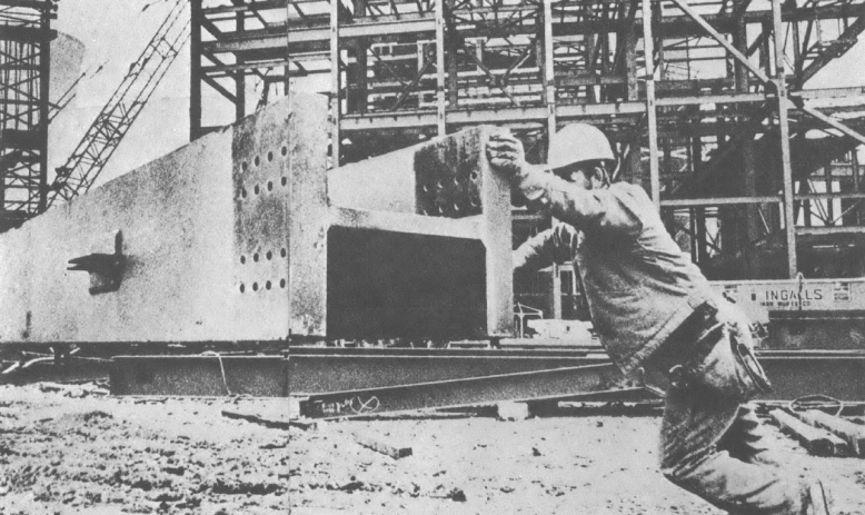 Black and white photo of man in hard hat pushing large steel beam across construction site