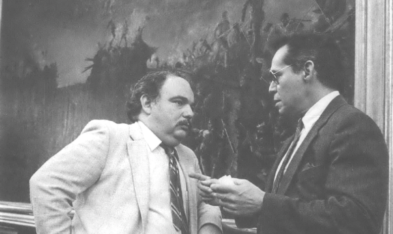 Photograph of two men in suits talking to each other in front of large landscape painting