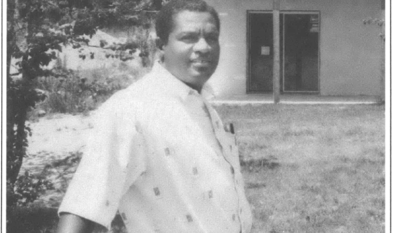 man standing on a porch leaning on railing in a white shirt backyard in a background