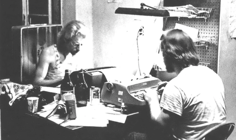 Two men at a desk, typing on typewriters