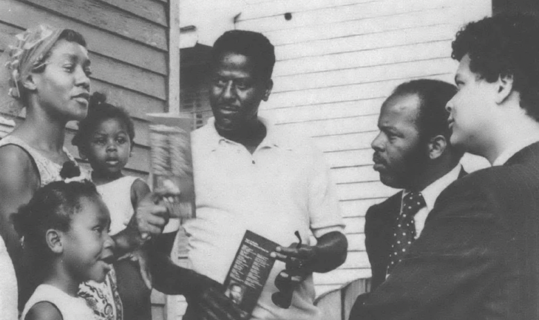 Black and white photo of Julian Bond and John Lewis talking with Black family outside a house