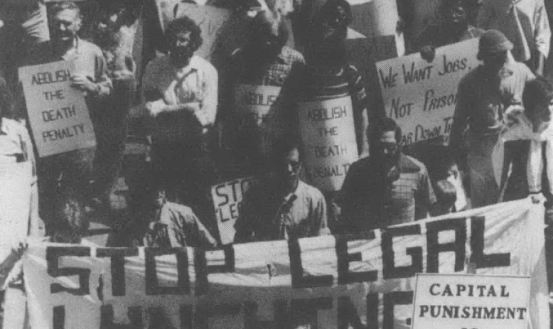Black and white aerial shot of group of people holding signs, including banner reading "Stop Legal Lynching"