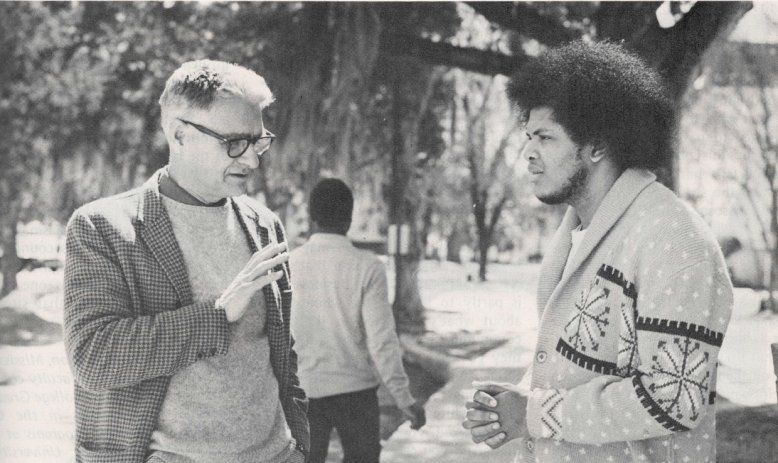 Black and white photo of white man in glasses talking to Black man in sweater outside under a tree
